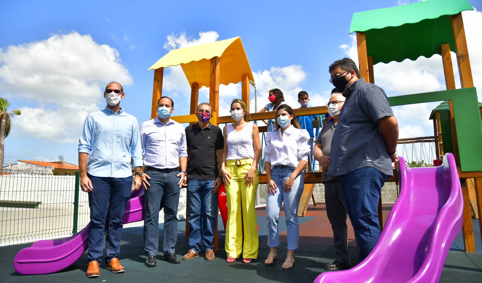 Carol Bezerra e Onélia Santana em pé em frente a parque infantil ao lado de 5 senhores e duas crianças ao fundo em cima do brinquedo